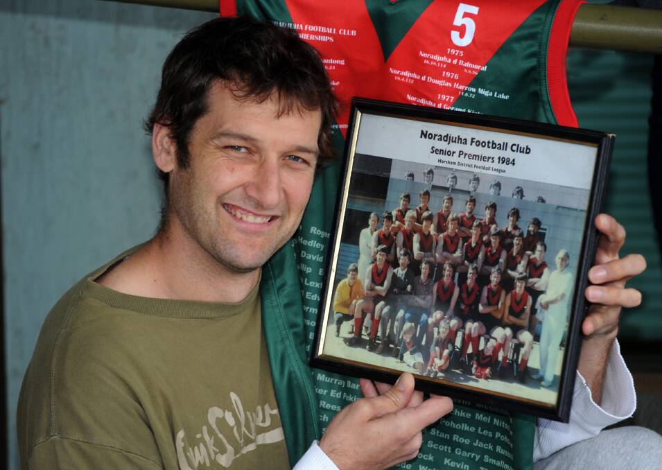 BACK TO THE FUTURE: Noradjuha-Quantong player Heath Watson with a picture of Noradjuha’s 1984 premiership side. He is one of the mascots in the foreground. Watson will play on Saturday in a game commemorating 30 years since the premiership. Behind Watson is one of the special jumpers senior players will wear to commemorate Noradjuha’s nine premierships. Picture: PAUL CARRACHER